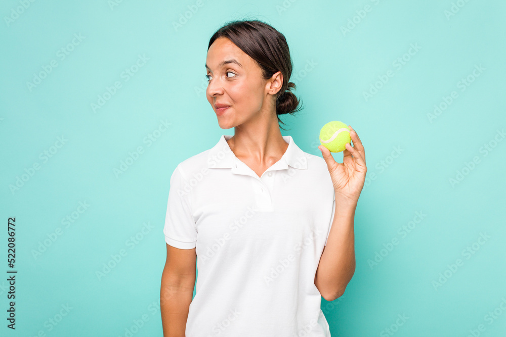 Young hispanic physiotherapy holding a tennis ball isolated on blue background looks aside smiling, cheerful and pleasant. Young hispanic physiotherapy holding a tennis ball isolated on blue background looks aside smiling, cheerful and pleasant.