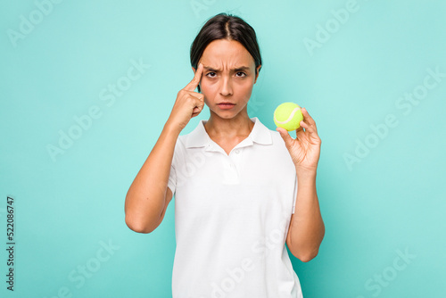Young hispanic physiotherapy holding a tennis ball isolated on blue background pointing temple with finger, thinking, focused on a task. © Asier