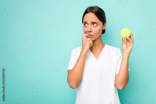 Young hispanic physiotherapy holding a tennis ball isolated on blue background relaxed thinking about something looking at a copy space. © Asier