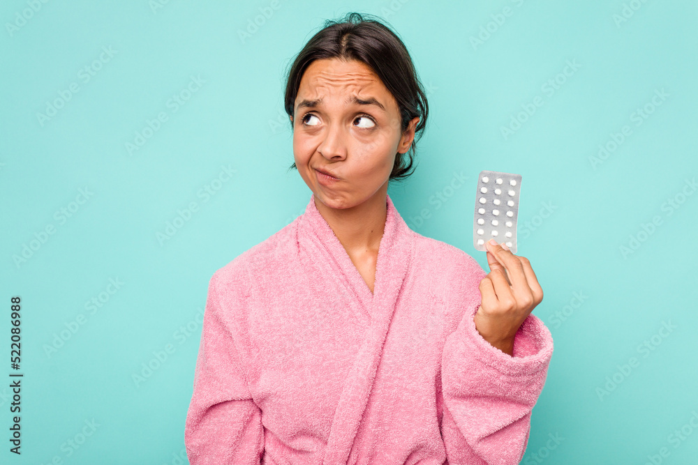Young hispanic woman holding pills isolated on blue background confused, feels doubtful and unsure. Young hispanic woman holding pills isolated on blue background confused, feels doubtful and unsure.