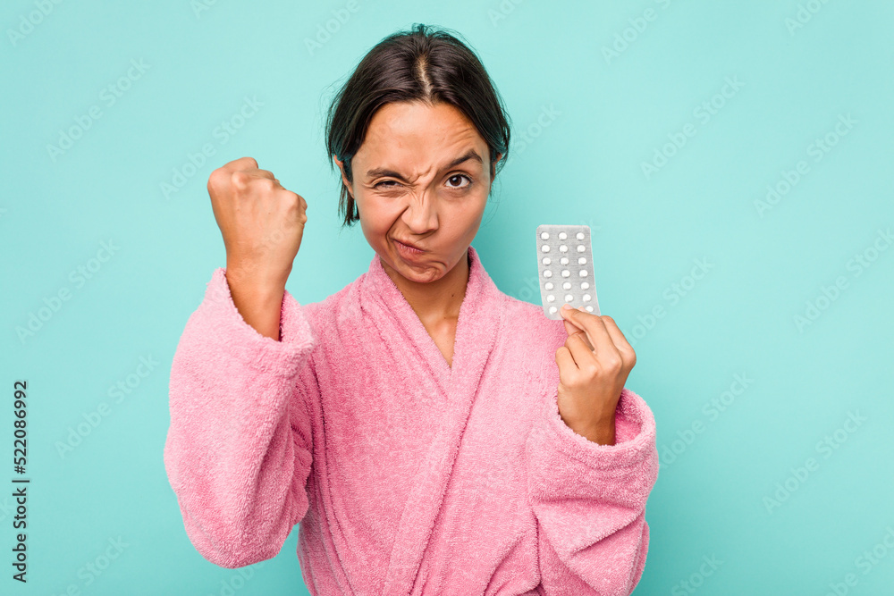 Young hispanic woman holding pills isolated on blue background showing fist to camera, aggressive facial expression. Young hispanic woman holding pills isolated on blue background showing fist to camera, aggressive facial expression.