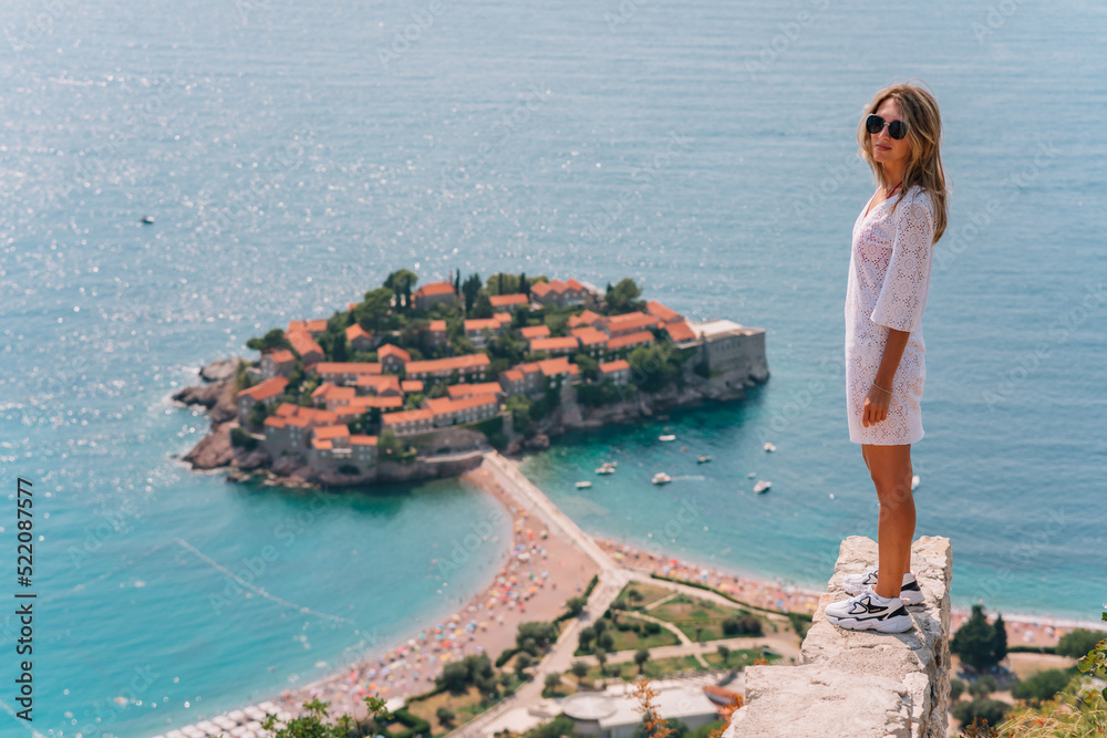 a girl poses on a mountain with a top view of the city of Sveti Stefan in Montenegro, a picturesque bay