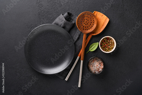 Cooking wooden utensils, black empty plate, basil leaves and spices on dark stone background. Abstract food background. Top view of dark rustic kitchen table with wooden cooking spoon, frame. Mockup.
