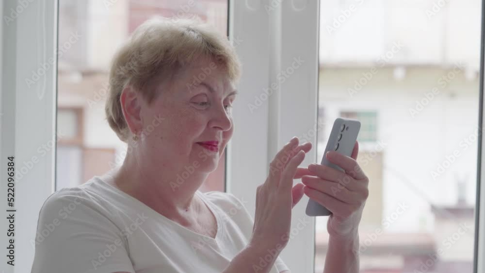 Older Middle Aged Blond Woman using and text messaging by mobile phone near Window with old town background, indoor, Relaxed mature old 60s woman, search ecommerce offers on cell phone technology devi Older Middle Aged Blond Woman using and text messaging by mobile phone near Window with old town background, indoor, Relaxed mature old 60s woman, search ecommerce offers on cell phone technology devi