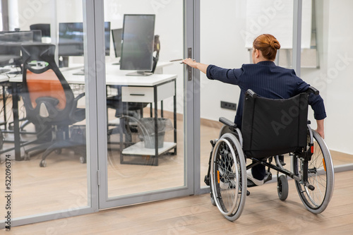 Red-haired caucasian woman in a wheelchair trying to open the door in the office. 