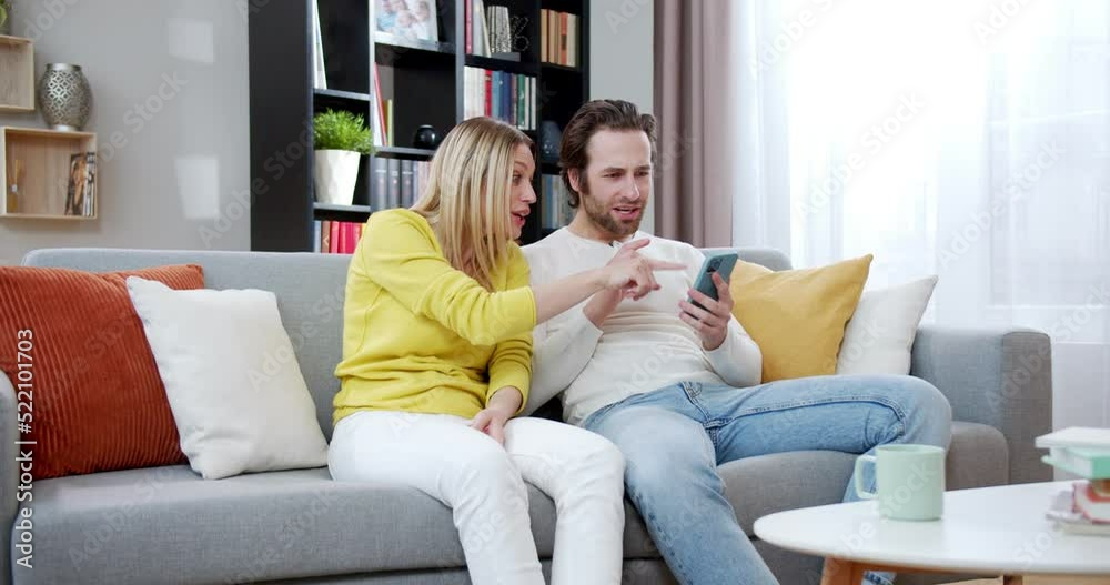 Cheerful Caucasian couple using smartphone together and laughing like they winning. At home on couch. Beautiful woman and handsome man watching something on mobile phone and cheering up. Cheerful Caucasian couple using smartphone together and laughing like they winning. At home on couch. Beautiful woman and handsome man watching something on mobile phone and cheering up.