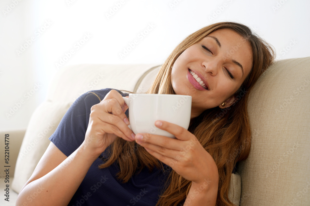Dreamy smiling positive girl eyes closed holds cup with warm beverage imagining sitting on sofa. Focus on woman face. Dreamy smiling positive girl eyes closed holds cup with warm beverage imagining sitting on sofa. Focus on woman face.