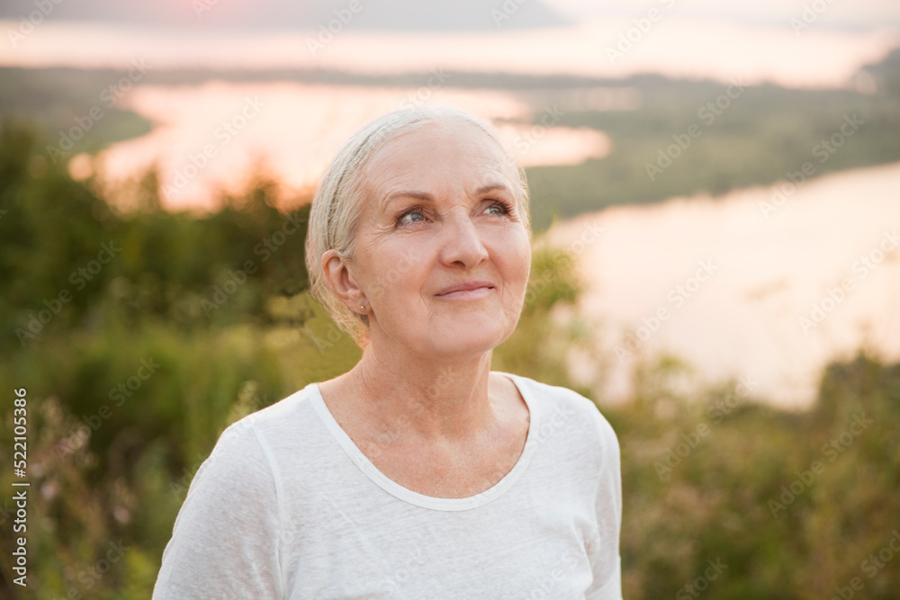 portrait of a smiling senior woman wearing a white T-shirt looks at the camera relaxed outdoors at sunset. copy space. Slow life. Enjoying the little things. spends time in nature in summer portrait of a smiling senior woman wearing a white T-shirt looks at the camera relaxed outdoors at sunset. copy space. Slow life. Enjoying the little things. spends time in nature in summer