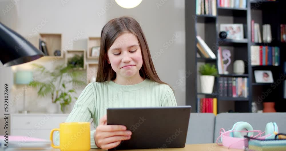 Cute Caucasian teen girl sitting at table in cozy living room and laughing while watching funny video on tablet device. At home. Beautiful happy teenage schoolgirl tapping and scrolling on computer. Cute Caucasian teen girl sitting at table in cozy living room and laughing while watching funny video on tablet device. At home. Beautiful happy teenage schoolgirl tapping and scrolling on computer.