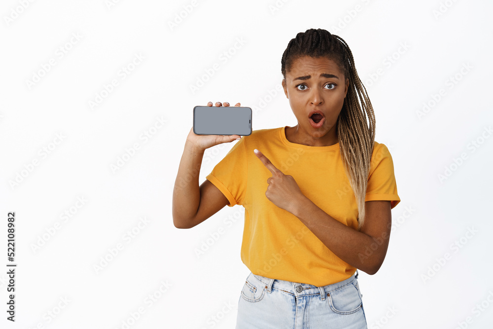 Shocked young Black woman looks with disbelief, pointing finger at smartphone empty screen, application interface, online promo on phone, white background Shocked young Black woman looks with disbelief, pointing finger at smartphone empty screen, application interface, online promo on phone, white background