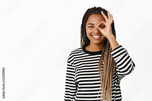 Portrait of smiling african woman shows okay, zero gesture on eye, looking happy, make approval sign, say yes, no problem, guarantee quality, white background © Cookie Studio