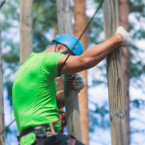 View of high ropes course, process of climbing in amusement acitivity rope park, passing obstacles and zip line on heights in climbing safety equipment gear between the trees, summer sunny day