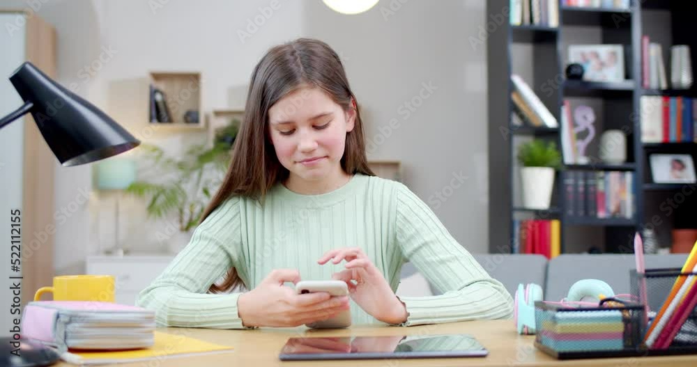 Pretty Caucasian teen cute girl texting and chatting on mobile phone while sitting at desk in cozy living room. Indoor. Beautiful happy smiled schoolgirl tapping and scrolling on smartphone. Pretty Caucasian teen cute girl texting and chatting on mobile phone while sitting at desk in cozy living room. Indoor. Beautiful happy smiled schoolgirl tapping and scrolling on smartphone.