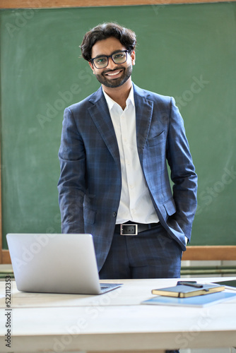 Smiling young indian business man manager wearing suit looking at camera standing in office. Arab teacher or professor posing for portrait at work desk with laptop in front of blackboard. Vertical photo