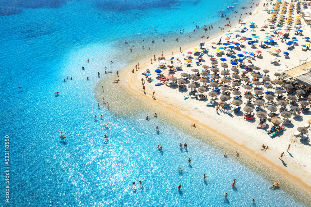 Aerial view of colorful umbrellas on sandy beach, people in blue sea at sunset in summer. Tuerredda Beach, Sardinia, Italy. Tropical landscape with turquoise water. Travel and vacation. Top view	