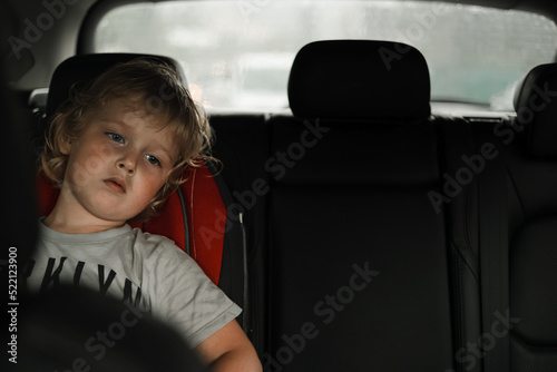 Thoughtful little blond baby boy seated in car backseat looking through rain covered window in rainy day.