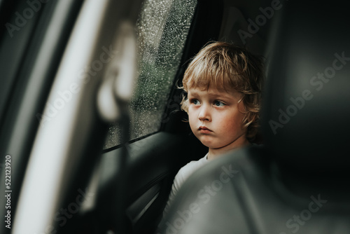 Thoughtful little blond baby boy seated in car backseat looking through rain covered window in rainy day.