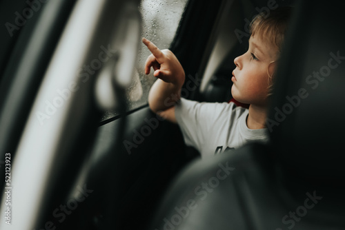 Thoughtful little blond baby boy seated in car backseat looking through rain covered window in rainy day.