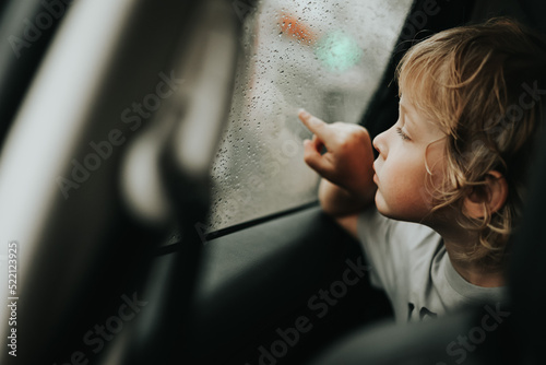 Thoughtful little blond baby boy seated in car backseat looking through rain covered window in rainy day.
