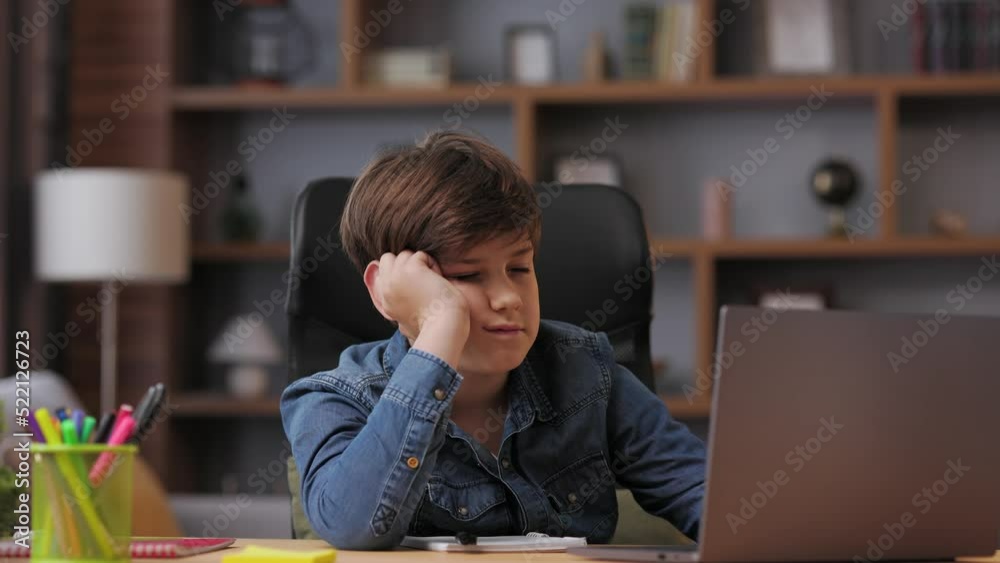 Young boy studying remotely at home using laptop. Tired schoolboy falling asleep during an online lesson with teacher. Distance learning is boring and uninteresting. Young boy studying remotely at home using laptop. Tired schoolboy falling asleep during an online lesson with teacher. Distance learning is boring and uninteresting.