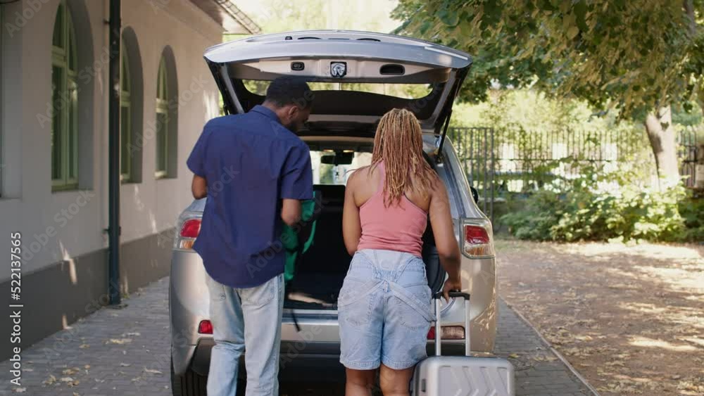 African american couple loading vehicle trunk with voyage luggage while getting ready for holiday citybreak. People putting baggage and trolleys inside car while getting ready for field trip departure