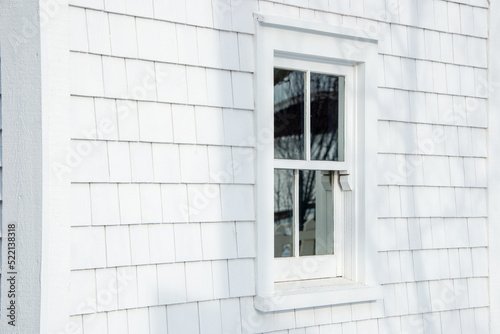 The exterior of a vintage building is covered in white cedar wood shingles. There's a small single hung glass window in the center with the reflection of a white picket fence and tree in the window.  © Dolores  Harvey