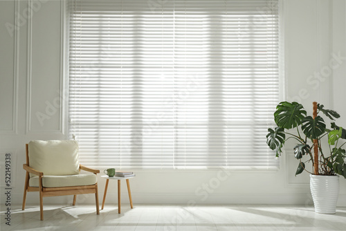 Cosy armchair and houseplant near large window with blinds in spacious room. Interior design