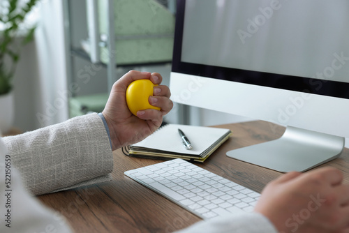 Man squeezing antistress ball while working with computer in office, closeup photo