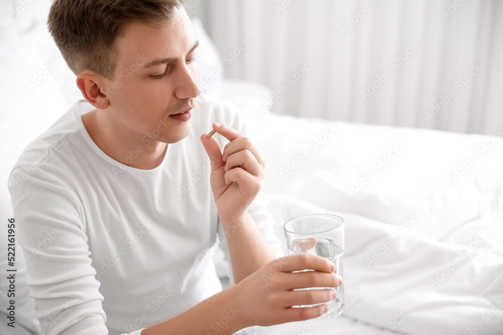 Young man with glass of water taking vitamin supplement in bedroom Young man with glass of water taking vitamin supplement in bedroom