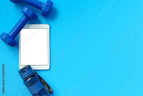 Modern tablet computer, dumbbells and bottle of water on blue background