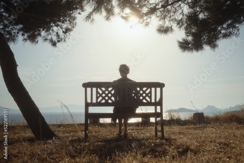 A woman in a dress sits on a bench and looks at the blue sea. photo
