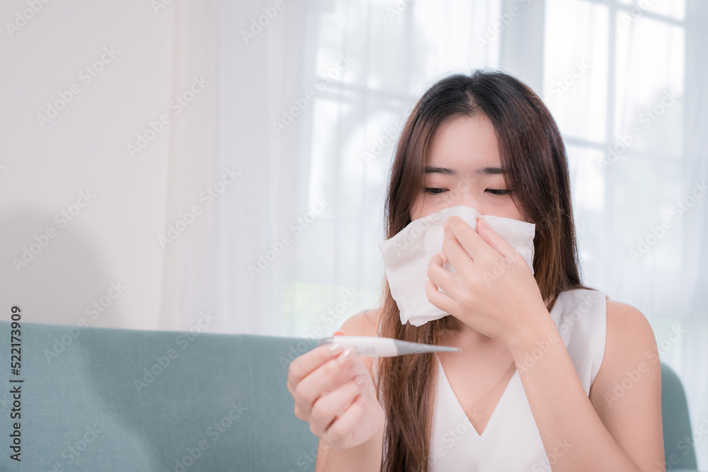 Asian woman patient with tissue her sick symptom in living room at home, Sick young female sitting under the blanket on sofa coughing or sneezing in tissue. Asian woman patient with tissue her sick symptom in living room at home, Sick young female sitting under the blanket on sofa coughing or sneezing in tissue.
