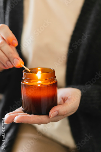 Woman holding in hand candle and light candle in small amber glass jar with wooden wick. Zen and relax concept