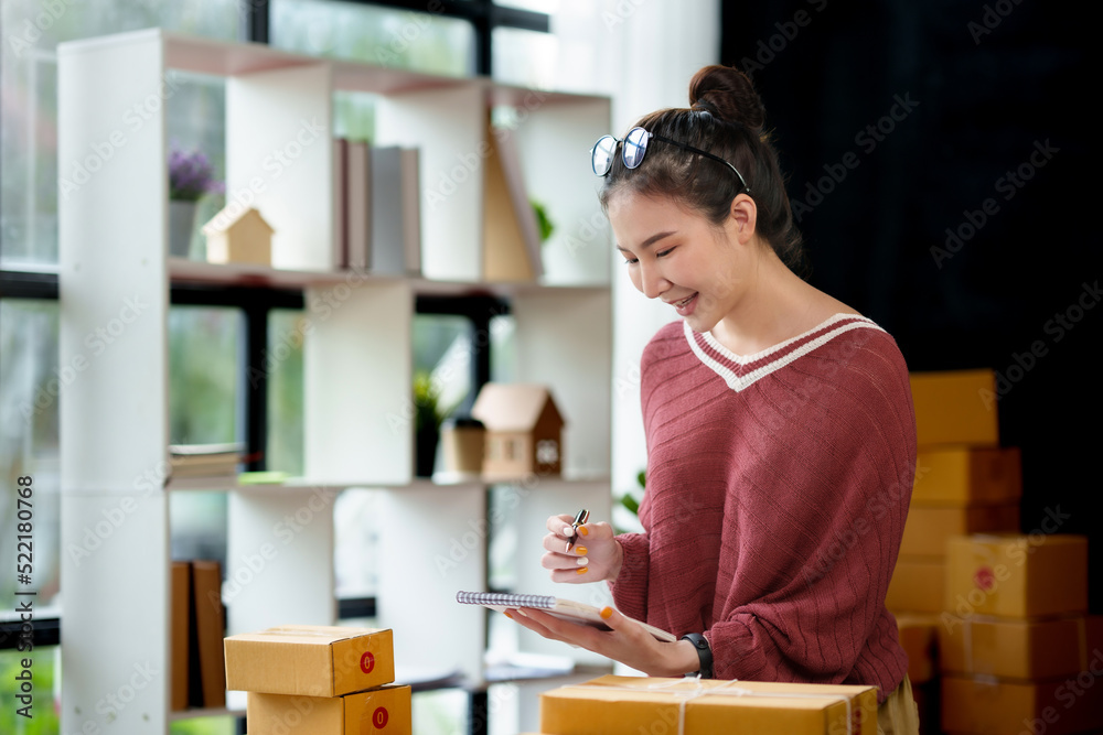 Young woman, an Asian online business owner, smiles, prepares parcel boxes and checks online orders for products to be delivered to customers with her laptop. Young woman, an Asian online business owner, smiles, prepares parcel boxes and checks online orders for products to be delivered to customers with her laptop.