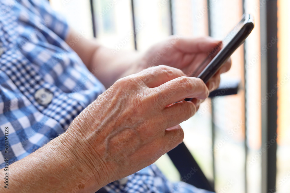 Elderly woman sitting with smartphone in chair, mobile phone in wrinkled female hands closeup. Concept of online communication in retirement