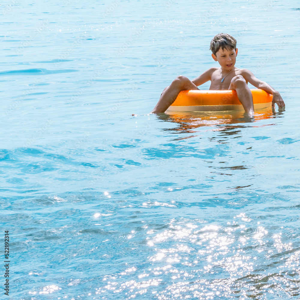 A pensive white Eastern European boy of 9 years old is riding a swim ring under the bright sun in the sea. Copy space for your text