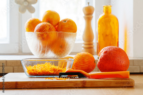 Oranges and orange zest prepared for confiture on a cutting board next to a curly paring knife against a background of peeled oranges in a bowl