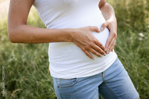 pretty attractive young pregnant woman with white top is standing in high meadow and forming a heart with her hands on her belly out in the nature