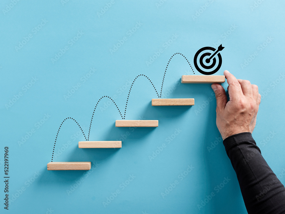 Male hand arranges a wooden block staircase with target icon. Achieving goals and objectives or goal setting - obrazy, fototapety, plakaty 
