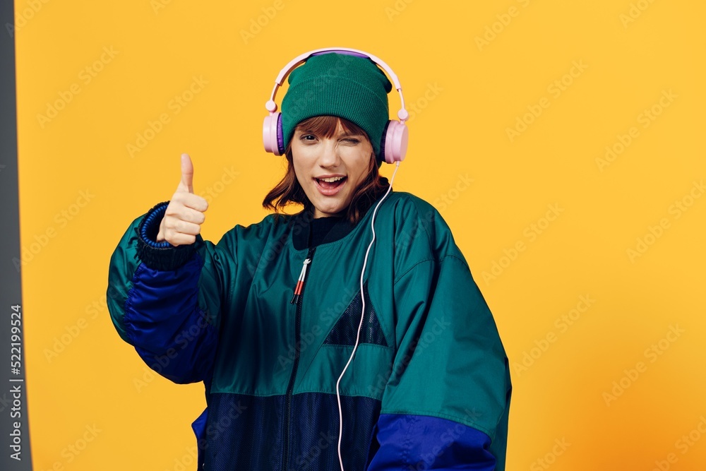 a joyful woman in a green jacket and a hat in the style of the 90s enjoys music standing in pink headphones on a yellow background showing a thumbs up