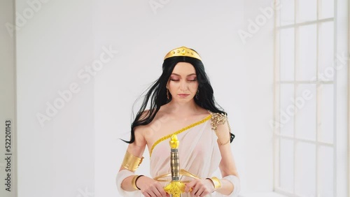 Fantasy portrait woman Greek goddess of war Athena holding ancient steel sword in hand. Creative design silk clothing white creative dress gold accessories crown vintage costume. Long hair fly in wind photo