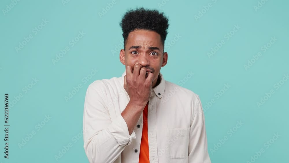 Young scared fearful man of African American ethnicity 20s he wear shirt t-shirt looking camera biting nails fingers isolated on plain pastel light blue cyan color background. People lifestyle concept Young scared fearful man of African American ethnicity 20s he wear shirt t-shirt looking camera biting nails fingers isolated on plain pastel light blue cyan color background. People lifestyle concept