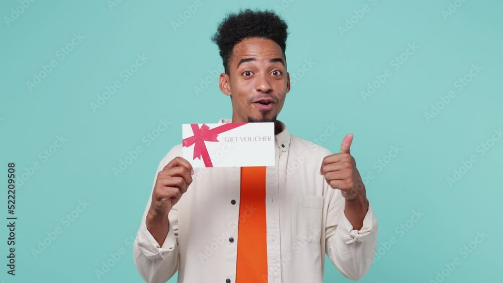 Young surprised shocked happy man of African American ethnicity wear shirt t-shirt point finger on store gift certificate voucher do winner gesture isolated on plain pastel light blue cyan background Young surprised shocked happy man of African American ethnicity wear shirt t-shirt point finger on store gift certificate voucher do winner gesture isolated on plain pastel light blue cyan background