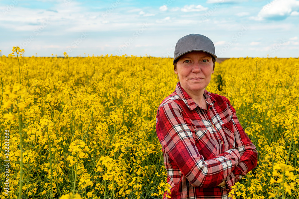Confident and self-assured farm worker wearing red plaid shirt and trucker's hat standing in cultivated rapeseed field in bloom and looking over crops
