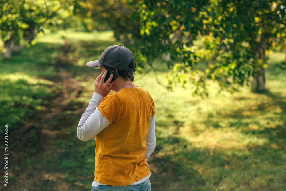 Female farmer using smartphone in walnut tree orchard. Farm worker wearing orange t-shirt and trucker's hat with mobile phone in hand. Female farmer using smartphone in walnut tree orchard. Farm worker wearing orange t-shirt and trucker's hat with mobile phone in hand.