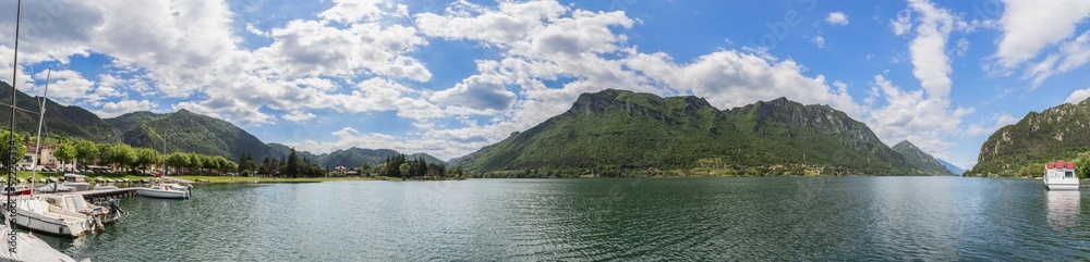 Maxi panoramic shot of glacier Lombard highland lake Idro (Lago d'Idro) surrounded by alpine wooded cliffs under piercing blue sky, several boats at a pier. Brescia, Lombardy, Italy