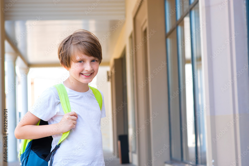 handsome happy little boy schoolboy with a backpack goes to school, stands at the school building handsome happy little boy schoolboy with a backpack goes to school, stands at the school building