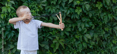 a boy in a T-shirt on the background of a hedge aims a slingshot. banner with space for text