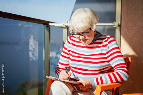 Elderly woman in glasses sitting on a balcony near the sea and looking at magazine, solving a crossword