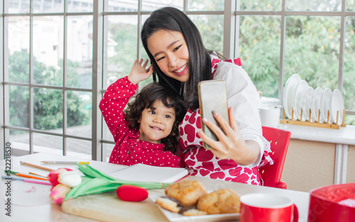 Beautiful Asian mother, adorable little daughter wearing casual clothes, sitting at home, playing, video conference, online studying, talking on phone, smiling with happiness. Kid, Education Concept.